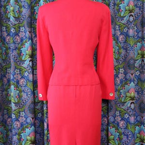 May include: A bright red two-piece suit, consisting of a long-sleeved jacket and a knee-length skirt. The jacket has silver buttons on the sleeves. The suit is displayed on a mannequin against a patterned blue curtain.