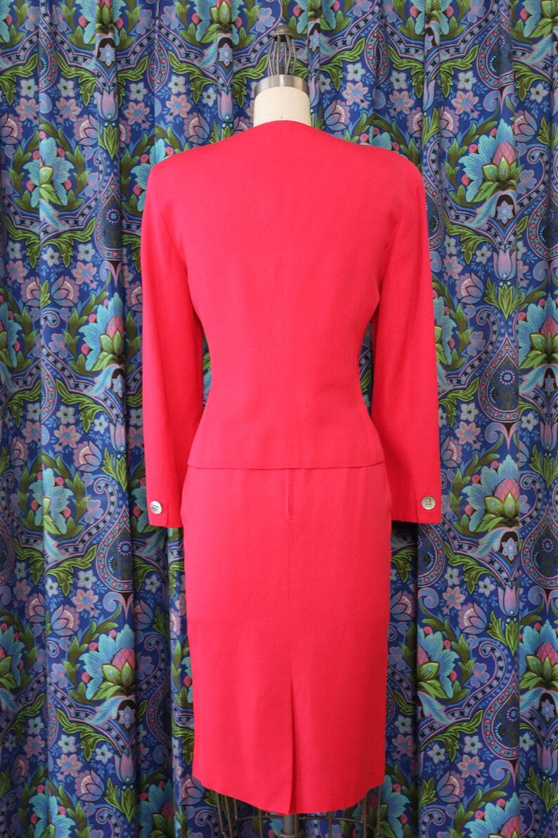 May include: A bright red two-piece suit, consisting of a long-sleeved jacket and a knee-length skirt. The jacket has silver buttons on the sleeves. The suit is displayed on a mannequin against a patterned blue curtain.