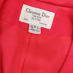May include: Close-up of a vibrant red garment, possibly a suit jacket or dress, with a label that reads "Christian Dior THE SUIT PETITES PARIS NEW YORK." The label also indicates a size 8 and "IMPORTED ORIGIN."
