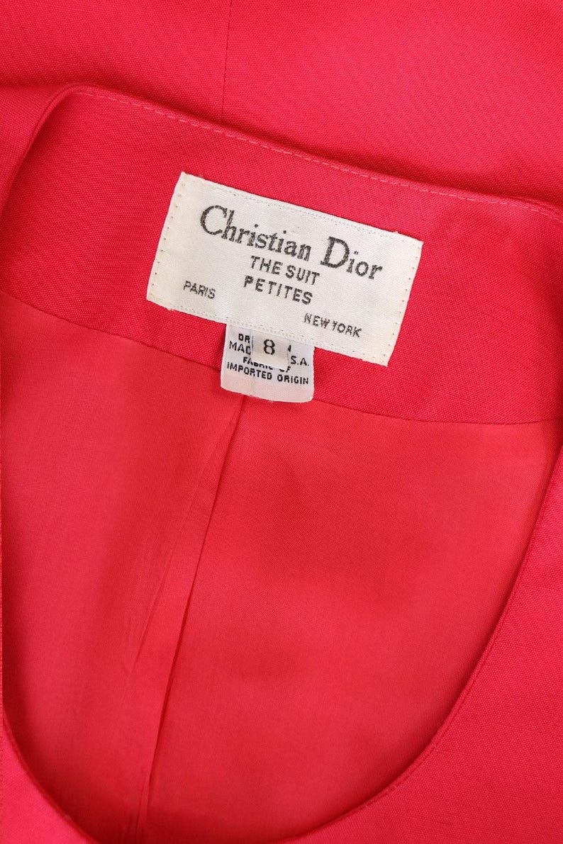 May include: Close-up of a vibrant red garment, possibly a suit jacket or dress, with a label that reads "Christian Dior THE SUIT PETITES PARIS NEW YORK." The label also indicates a size 8 and "IMPORTED ORIGIN."