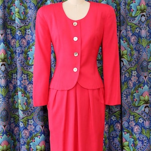 May include: A bright pink vintage suit, consisting of a long-sleeved jacket with three buttons and a matching knee-length skirt. The jacket has a rounded neckline and a peplum-style waist. The suit is displayed on a mannequin against a patterned blue background.