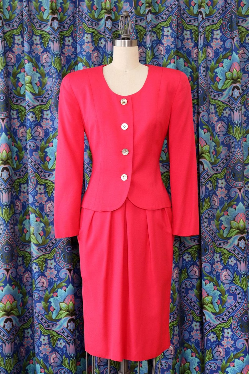May include: A bright pink vintage suit, consisting of a long-sleeved jacket with three buttons and a matching knee-length skirt. The jacket has a rounded neckline and a peplum-style waist. The suit is displayed on a mannequin against a patterned blue background.