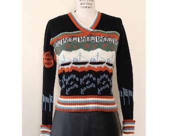 May include: A black and white knit sweater with a colorful geometric pattern. The sweater features a V-neck, long sleeves, and a striped waistband. The pattern includes ships, flags, and the text "Levante a River".