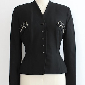 May include: Black blazer with a V-neck and six buttons. The blazer has a tailored fit and features decorative accents on the chest. The sleeves are long with folded cuffs. The jacket is displayed on a mannequin.
