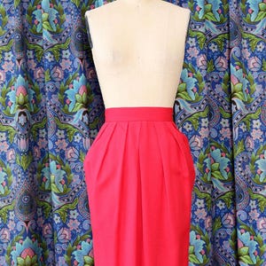 May include: A bright pink pencil skirt with pleats and pockets, displayed on a mannequin. The skirt is knee-length and has a high waist. The background features a blue floral patterned fabric.