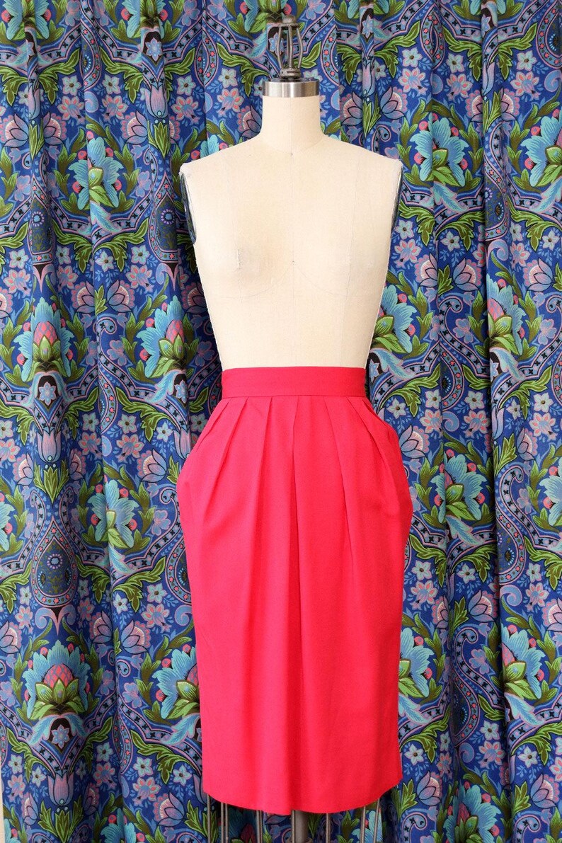 May include: A bright pink pencil skirt with pleats and pockets, displayed on a mannequin. The skirt is knee-length and has a high waist. The background features a blue floral patterned fabric.