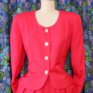 May include: A bright red, two-piece suit displayed on a mannequin. The jacket has a rounded neckline, long sleeves, and white buttons. The skirt is pleated. The background features a blue floral patterned curtain.