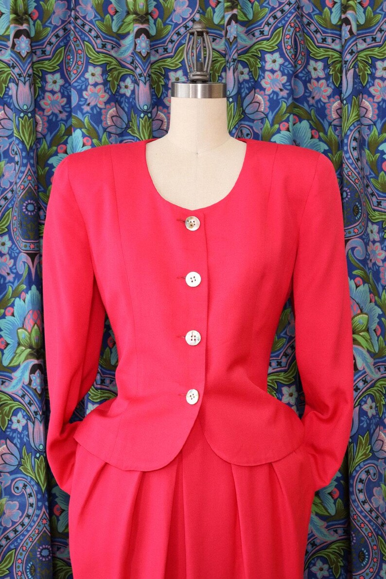 May include: A bright red, two-piece suit displayed on a mannequin. The jacket has a rounded neckline, long sleeves, and white buttons. The skirt is pleated. The background features a blue floral patterned curtain.
