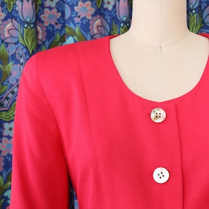 May include: A bright red blazer with a round neckline and long sleeves. The blazer features three white, decorative buttons down the front. The garment is displayed on a mannequin against a patterned blue background.