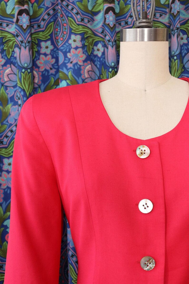 May include: A bright red blazer with a round neckline and long sleeves. The blazer features three white, decorative buttons down the front. The garment is displayed on a mannequin against a patterned blue background.