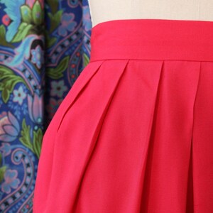 May include: Close-up of a vibrant red skirt with pleats. The skirt has a wide waistband and a tailored design. The fabric appears smooth and has a slight sheen. The background features a blue floral patterned fabric.
