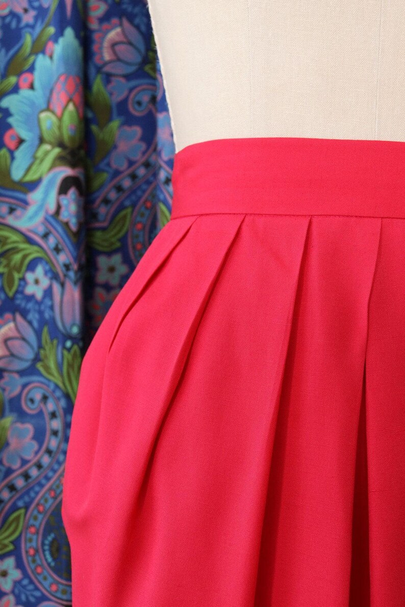 May include: Close-up of a vibrant red skirt with pleats. The skirt has a wide waistband and a tailored design. The fabric appears smooth and has a slight sheen. The background features a blue floral patterned fabric.