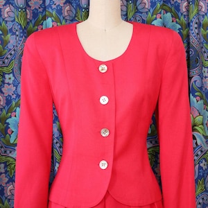 May include: A bright red, two-piece suit displayed on a mannequin. The jacket has a rounded neckline, long sleeves, and is fastened with four white buttons. The skirt is pleated. The background features a blue floral pattern.
