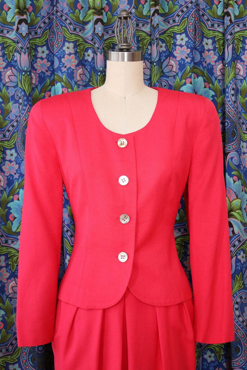 May include: A bright red, two-piece suit displayed on a mannequin. The jacket has a rounded neckline, long sleeves, and is fastened with four white buttons. The skirt is pleated. The background features a blue floral pattern.