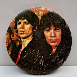 May include: Circular button with a vintage image of two men with long hair and dark jackets. The button has a retro aesthetic, with a slightly faded appearance. The image is set against a neutral background.