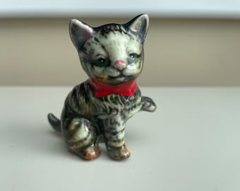 May include: A ceramic figurine of a seated kitten wearing a red bow tie. The cat has green eyes, a pink nose, and a black and grey striped coat. The figurine is a decorative item.