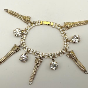 May include: Gold-coloured bracelet adorned with clear rhinestones. The bracelet has a double-strand design with dangling charms and decorative elements. The charms include large, round rhinestones and elongated, cone-shaped accents, all encrusted with sparkling stones.