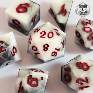 May include: A selection of polyhedral dice featuring a marbled white and grey design. The numbers are painted in a vibrant red. The dice are in various shapes, including a 20-sided die. The Souls & Stuff logo is visible in the upper right corner.