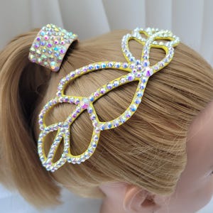 May include: A yellow hair accessory featuring an intricate, open-work leaf-like design completely covered in iridescent, multi-faceted crystals. A matching cylindrical hair tie, also adorned with the same sparkling crystals, sits above.