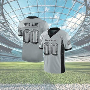 Puede incluir: Dos camisetas de f&uacute;tbol grises con detalles negros y el n&uacute;mero "00". Una camiseta muestra "YOUR NAME" y la otra "TEAM NAME". Las camisetas est&aacute;n sobre un campo verde con un fondo de estadio.