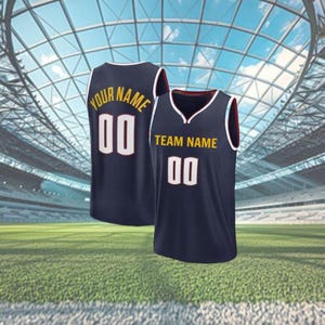 May include: Two navy blue basketball vests with white and gold lettering and numbers. One vest reads "YOUR NAME" and "00", the other reads "TEAM NAME" and "00". The vests have white trim and are set against a stadium background.