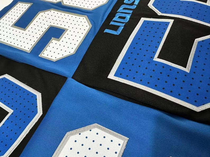 Puede incluir: Primer plano de camisetas deportivas azules y negras. Las camisetas presentan grandes n&uacute;meros blancos con un patr&oacute;n de puntos y ribetes plateados. La palabra "Lions" es visible en letras azules en una camiseta negra.