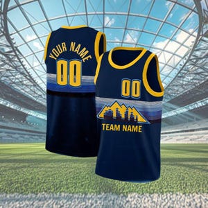 May include: Two navy blue basketball vests with yellow trim. One vest has "YOUR NAME" and "00" on the back. The other vest has "00" and "TEAM NAME" with a mountain graphic on the front. Both vests have a blue, white, and yellow striped design.