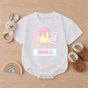 May include: White baby onesie with a colorful popsicle design and the name "Amelia". The design features pink, yellow, and purple popsicles. A wooden bunny rattle and brown baby shoes are also in the image.