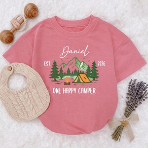 May include: A dusty rose-colored baby bodysuit with a camping-themed graphic. The graphic includes a mountain range, trees, a campfire, and a tent. The text reads "Daniel EST. 2026 ONE HAPPY CAMPER."