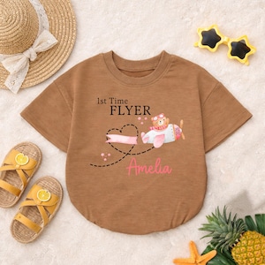 May include: Brown baby bodysuit with the text "1st Time FLYER" and the name "Amelia". The graphic features a bear in a plane with pink accents. The bodysuit is surrounded by summer accessories.