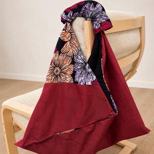 Può includere: Una borsa in tessuto con una sezione in tinta unita rosso scuro e un pannello a contrasto su fondo nero stampato con grandi motivi floreali arancioni e viola. La borsa presenta un manico annodato e una forma triangolare con cuciture.