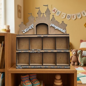 Può includere: Uno scaffale espositivo in legno marrone chiaro a forma di castello con scomparti quadrati. La parte superiore riporta le scritte Disney e Playmobil. Sotto, le etichette includono Mickey, Donald, Genie, Aladdin, Fee, Cupchette, Peter Pan, Vaiana, Maui, Woody, Jessie, Wdes, Triactible, Elastigirl, Pinocchio, Gepetto, Hector, Miguel, Hercule e Megara.