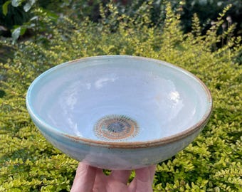 May include: A handmade ceramic bowl with a light blue interior and a brown rim. The bowl has a decorative pattern in the center. The bowl is held in a hand against a backdrop of green foliage.