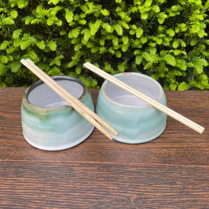 May include: Two small, handmade ceramic bowls with a light teal glaze and a white interior. Each bowl has a pair of wooden chopsticks resting on top. The bowls are set on a dark wooden surface, with green foliage in the background.