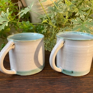 May include: Two handmade ceramic mugs. The mugs are white with a light blue interior and a matching blue accent at the base. Each mug has a curved handle. The mugs are displayed on a wooden surface.