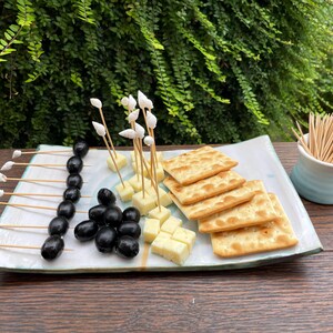 Puede incluir: Una bandeja rectangular de cerámica contiene aceitunas negras, cubos de queso amarillo y galletas cuadradas. Varias aceitunas están ensartadas en palillos de madera rematados con pequeñas conchas blancas.