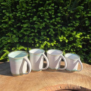 Puede incluir: Cuatro tazas de cerámica hechas a mano con un esmalte blanco y un interior y borde verde claro. Las tazas tienen una forma simple y redondeada con un asa grande. Se exhiben sobre una superficie de madera con un fondo verde.
