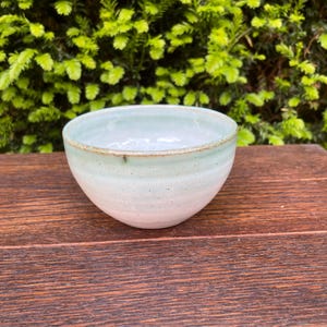 May include: A handmade ceramic bowl with a light green and white glaze. The bowl has a rounded shape and a slightly textured surface. The rim is a darker shade of green. The bowl is sitting on a wooden surface.