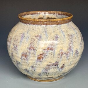 May include: A round ceramic vase with a wide opening and a textured surface. The vase features a cream-colored base with brown and purple accents. The rim is a darker brown. A decorative piece for the home.