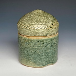 May include: A ceramic jar with a lid, featuring a green glaze with a speckled texture. The lid has a raised, textured design and a light brown glaze.