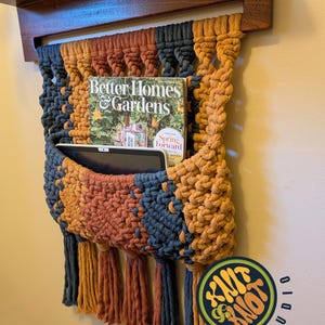 May include: A macrame wall hanging organizer featuring a dark wood dowel and alternating vertical stripes of navy blue, mustard yellow, and terracotta cord. The woven pocket holds a Better Homes & Gardens magazine and a tablet, with fringe at the bottom.