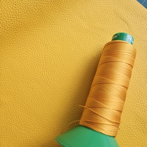 May include: A spool of golden-yellow thread rests on a textured, mustard-yellow leather swatch. The thread is wound around a green plastic spool. The leather has a pebbled surface, and the thread is likely for sewing or crafting.