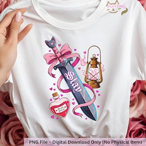May include: White t-shirt featuring a fantasy-themed graphic. The design includes a sword with a pink bow, a lantern, and a potion bottle. The word "Slay" is written on the sword. Small pink hearts are scattered around the design.