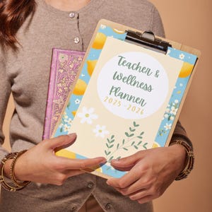 May include: A person holding a yellow and blue planner with lemons and flowers on the cover. The planner is titled "Teacher & Wellness Planner 2025-2026".