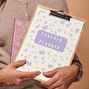 May include: A white clipboard with a floral pattern and the text "TEACHER PLANNER" in a purple box. The clipboard is held by a person wearing a brown sweater and bracelets.