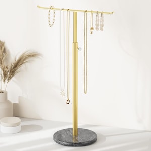 May include: A gold-colored jewelry display stand with a marble base. The stand holds various necklaces, bracelets, and earrings. The jewelry stand is adjustable and has a horizontal bar for hanging jewelry. The base is a dark gray color.