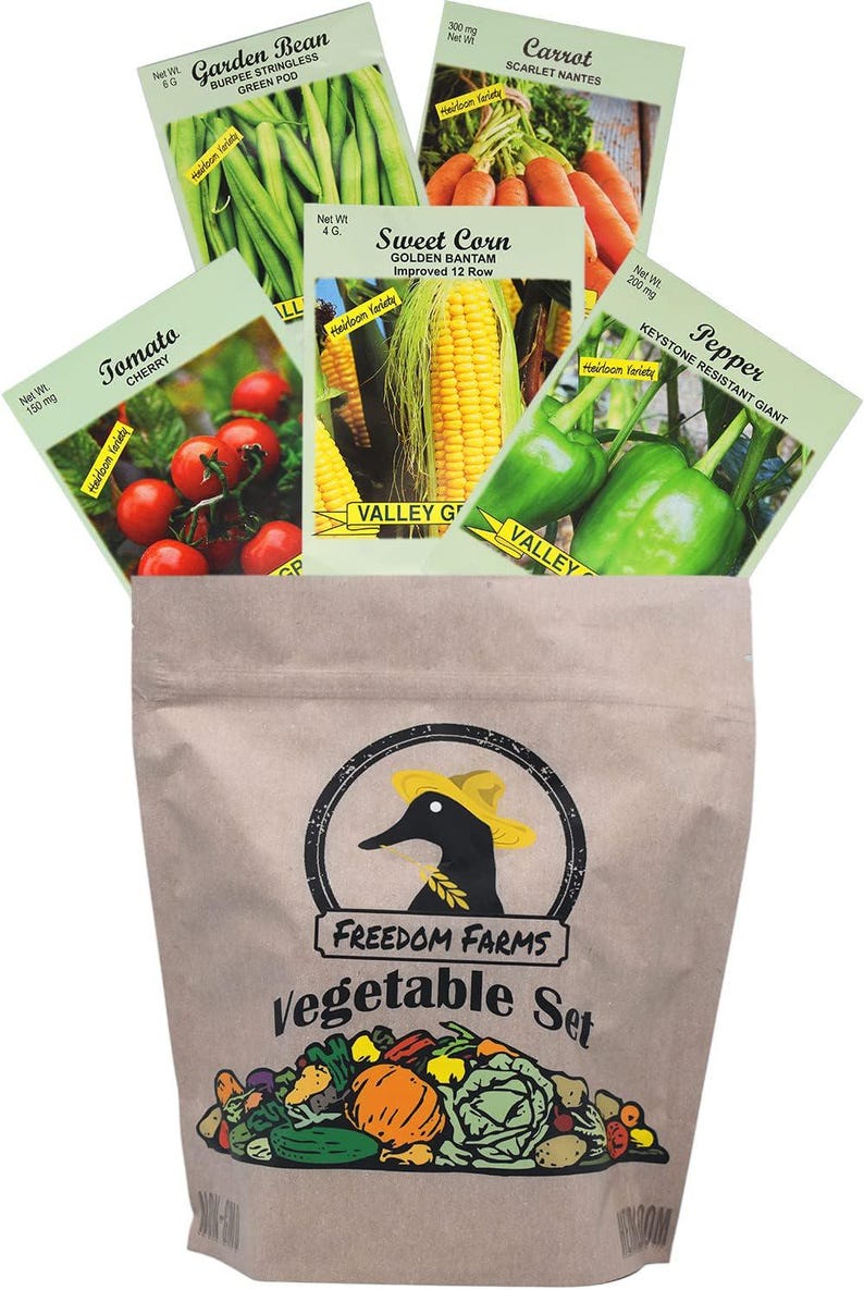 Op de afbeelding: Een bruine papieren zak met het label Freedom Farms Vegetable Set en een zwart eendenlogo, met vijf zakjes zaden: tuinboon, wortel, suikerma&iuml;s, tomaat en paprika. Elk zakje toont een kleurenfoto van de specifieke groentesoort.