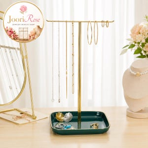 May include: A gold-toned metal jewelry stand with a T-shaped bar for hanging necklaces and earrings, featuring a rectangular dark green ceramic tray base for storing small accessories. The stand is displayed with various gold chains and hoops.