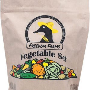 Op de afbeelding: Een beige papieren zakje met het label Freedom Farms Vegetable Set. Het logo toont een zwarte eend met een gele hoed die een graanstengel vasthoudt. Onder de tekst staat een kleurrijke grafische illustratie van diverse groenten zoals pompoen.