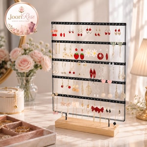 May include: A black metal jewelry display stand with five horizontal bars for hanging earrings, mounted on a rectangular light-colored wooden base. The stand is filled with various dangling earrings featuring diverse shapes, colors, and designs.